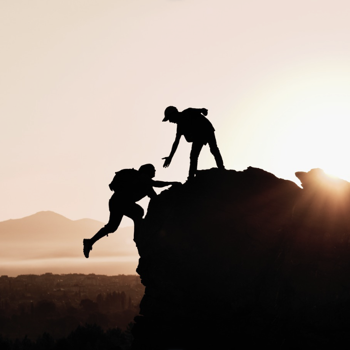 Deux randonneurs en silhouette, l'un aidant l'autre à grimper une roche au coucher du soleil.
