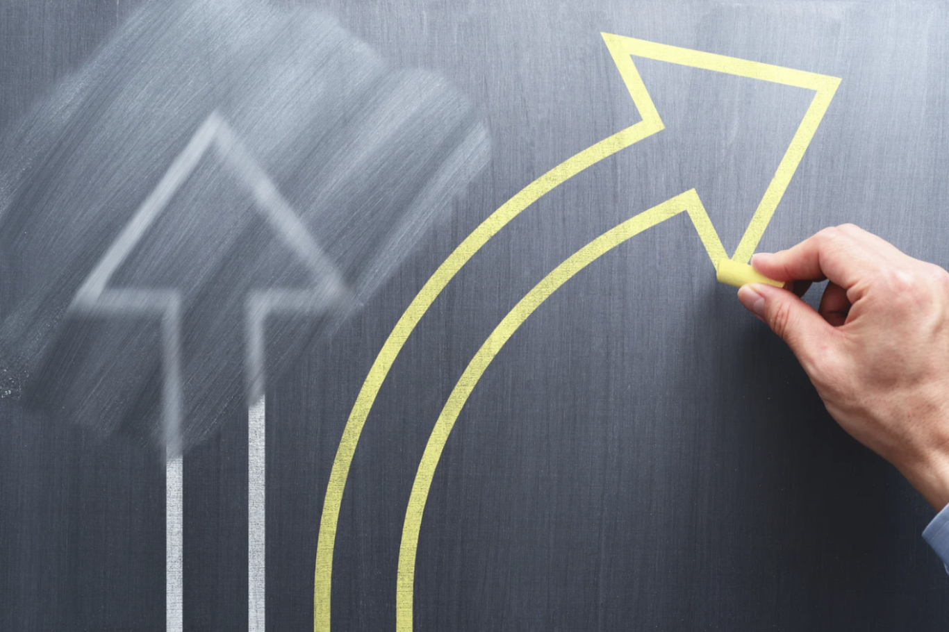 Main d’un homme traçant une nouvelle flèche jaune tournant sur un tableau noir, avec une flèche grise effacée en arrière-plan.